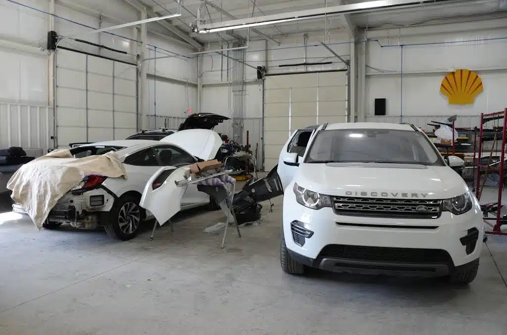An auto body repair shop with two vehicles in the workshop, including a white Land Rover Discovery and a car with a covered rear section being worked on, surrounded by tools and equipment.