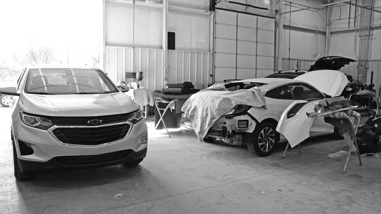 An automotive repair garage with a Chevrolet SUV parked on the left and a sedan undergoing bodywork on the right, covered partially with a protective cloth. The workspace features open tools and equipment in the background